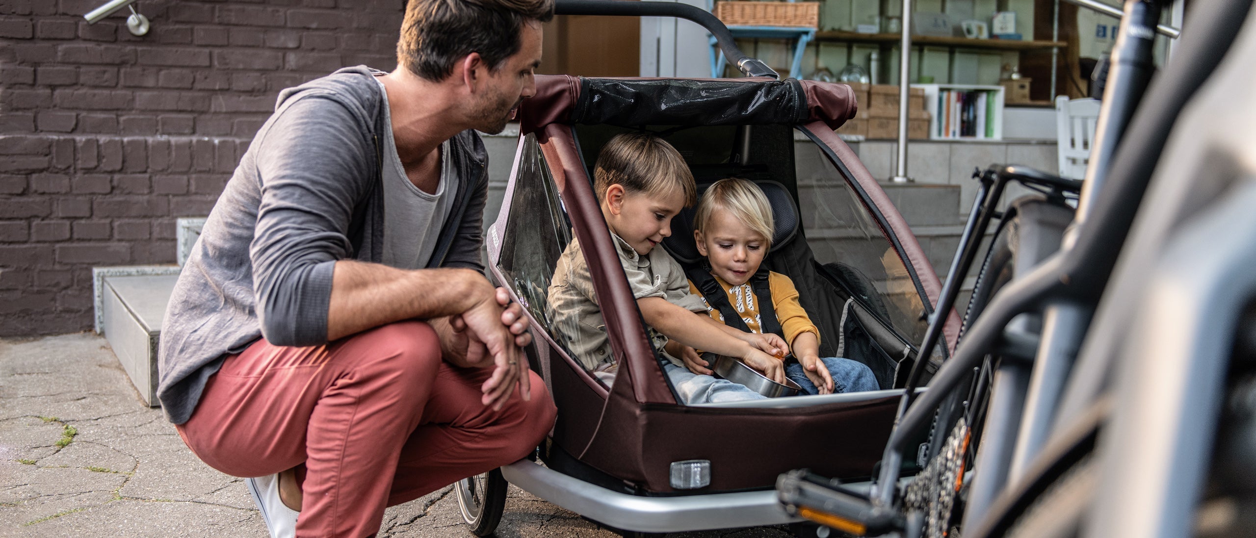 Two children sit in a Croozer child trailer eating gummy bears; dad crouches next to them, joyful atmosphere.